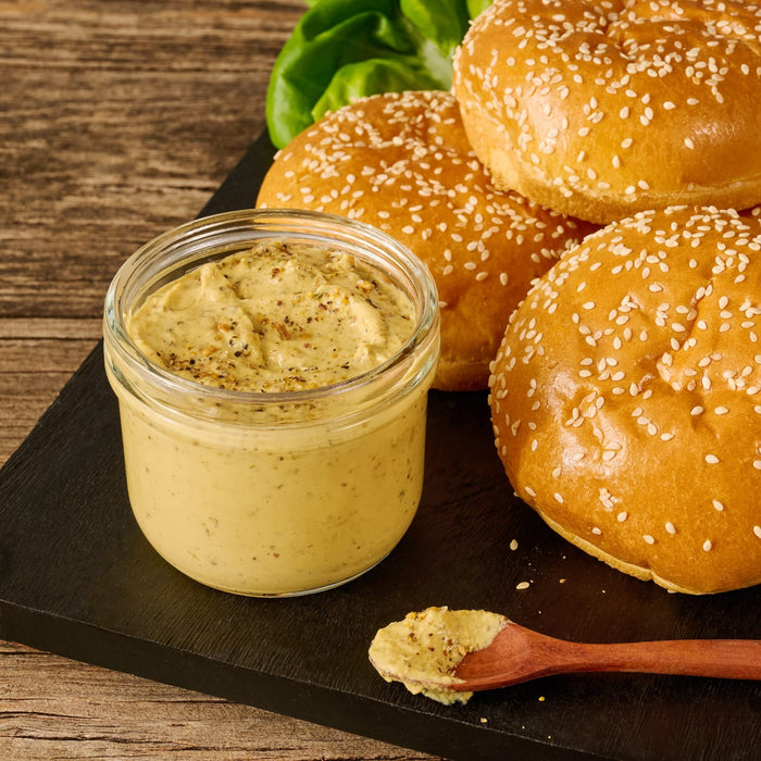 A creamy burger sauce, created using Mccormick Grill Mates Peppercorn And Worcestershire Seasoning Cracked 2.75 Oz, is presented in a glass jar alongside fresh sesame seed buns on a rustic wooden surface.