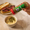 A hand sprinkles Mccormick Chimichurri Flavored Seasoning, 2.47 Oz, into a bowl of creamy aioli to be served with a steak sandwich in the background.