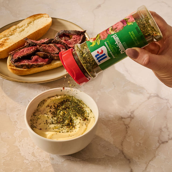 A hand sprinkles Mccormick Chimichurri Flavored Seasoning, 2.47 Oz, into a bowl of creamy aioli to be served with a steak sandwich in the background.