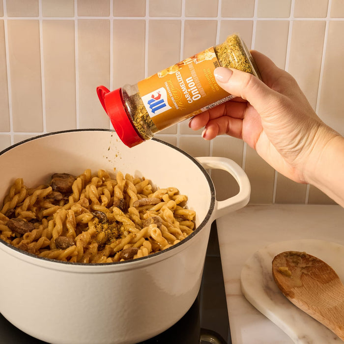 A hand sprinkles Mccormick Caramelized Onion Seasoning, 4.66 Oz, from its signature bottle over a creamy pasta and meat dish in a white pot.