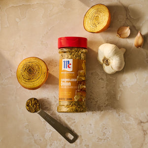 A jar of McCormick Signature Seasonings Caramelized Onion seasoning is displayed with sliced onions, garlic, and a measuring spoon filled with the seasoning.