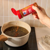 A hand sprinkles Mccormick Chili Garlic Seasoning, 5.22 Oz from a clear bottle into a light-colored pot filled with a dark liquid, adding flavor to a recipe.