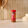 A bottle of Mccormick Chili Garlic Seasoning, 5.22 Oz, is centered on a kitchen counter, with a utensil holder and stacked plates visible in the background.