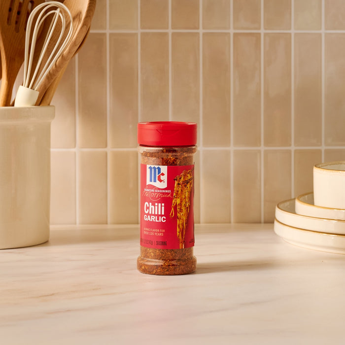 A bottle of Mccormick Chili Garlic Seasoning, 5.22 Oz, is centered on a kitchen counter, with a utensil holder and stacked plates visible in the background.