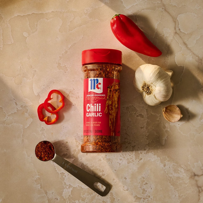 An overhead view shows a bottle of Mccormick Chili Garlic Seasoning, 5.22 Oz, surrounded by a fresh red chili pepper, a bulb of garlic, and a measuring spoon filled with the seasoning.