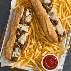 Two Philly cheesesteak sandwiches, with steak prepared using Mccormick Grill Mates Garlic Butter Seasoning, 6.56 Oz, are served on a bed of french fries with ketchup.