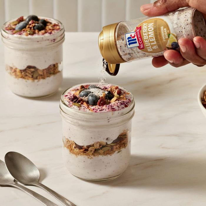 A hand sprinkles Mccormick Blueberry And Lemon Finishing Sugar, 3.43 Oz over a layered yogurt and granola parfait in a glass jar, adding a flavorful and decorative topping.