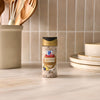 A bottle of Mccormick Blueberry And Lemon Finishing Sugar, 3.43 Oz, is centered on a marble kitchen countertop, with a utensil holder and plates in the background.