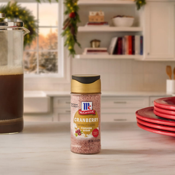 A bottle of Mccormick Cranberry Finishing Sugar, 3.43 Oz sits on a white kitchen counter in a festive holiday setting with a French press and red plates.