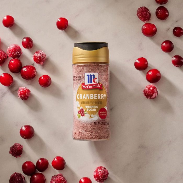 A bottle of Mccormick Cranberry Finishing Sugar, 3.43 Oz is centered on a marble surface surrounded by fresh and sugared cranberries.