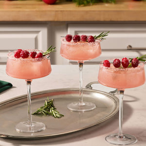 Three festive pink cocktails are served in coupe glasses with rims coated in Mccormick Cranberry Finishing Sugar, 3.43 Oz and garnished with sugared cranberries and rosemary sprigs.