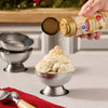 A hand sprinkles Mccormick Sugar Cookie Finishing Sugar, 3.39 Oz onto a tall scoop of vanilla ice cream served in a silver metal bowl.