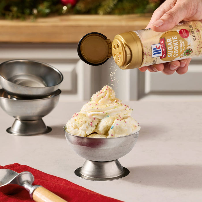 A hand sprinkles Mccormick Sugar Cookie Finishing Sugar, 3.39 Oz onto a tall scoop of vanilla ice cream served in a silver metal bowl.