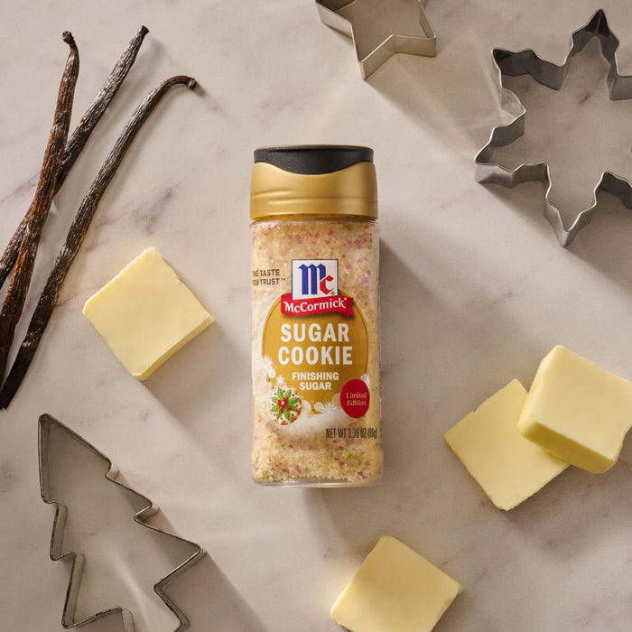 A bottle of Mccormick Sugar Cookie Finishing Sugar, 3.39 Oz is centered on a marble surface surrounded by festive baking essentials like vanilla beans, pats of butter, and holiday cookie cutters.