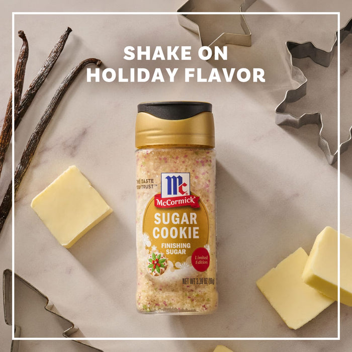 A bottle of Mccormick Sugar Cookie Finishing Sugar, 3.39 Oz is centered on a marble surface surrounded by vanilla beans, butter pats, and holiday cookie cutters under the text Shake On Holiday Flavor.