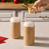 A hand sprinkles Mccormick Dulce De Leche Finishing Sugar, 3.43 Oz onto the thick foam of two iced coffee drinks in a festive kitchen setting.