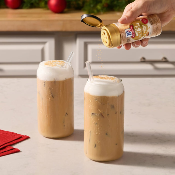 A hand sprinkles Mccormick Dulce De Leche Finishing Sugar, 3.43 Oz onto the thick foam of two iced coffee drinks in a festive kitchen setting.