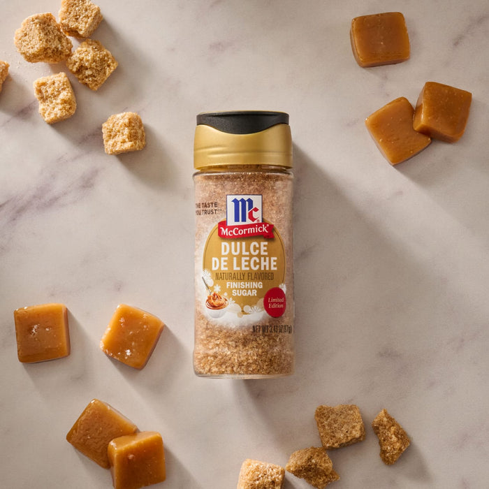 A bottle of Mccormick Dulce De Leche Finishing Sugar, 3.43 Oz is centered on a marble surface surrounded by square caramel candies and brown sugar cubes.