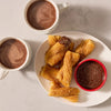 A plate of golden brown churros is generously coated with Mccormick Dulce De Leche Finishing Sugar 3.43 Oz and served alongside two mugs of hot chocolate and a small dipping bowl of chocolate sauce.