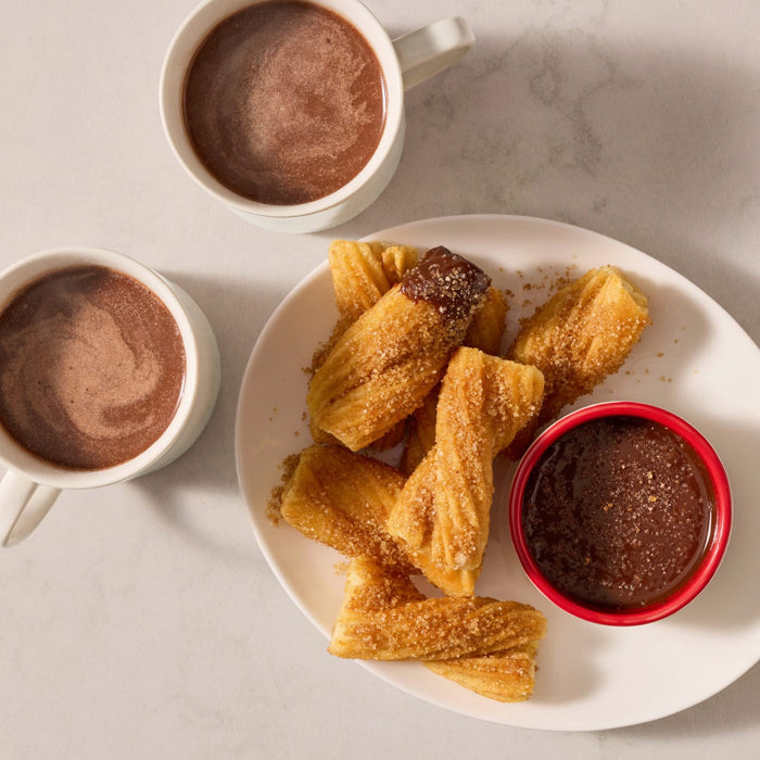 A plate of golden brown churros is generously coated with Mccormick Dulce De Leche Finishing Sugar 3.43 Oz and served alongside two mugs of hot chocolate and a small dipping bowl of chocolate sauce.