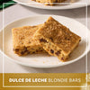 Two thick dulce de leche blondie bars with chocolate chips are served on a white plate and topped with a generous dusting of Mccormick Dulce De Leche Finishing Sugar, 3.43 Oz.