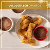 A plate of golden brown churros and a small bowl of dipping sauce are generously coated with Mccormick Dulce De Leche Finishing Sugar, 3.43 Oz, served alongside two mugs of hot chocolate.