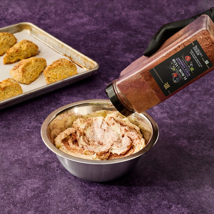A gloved hand sprinkles McCormick 2026 Flavor of the Year Sweet &amp; Smoky Naturally Flavored Black Currant Seasoning into a bowl of cream, with scones on a baking sheet in the background.