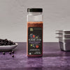 A bottle of McCormick 2026 Flavor of the Year Sweet &amp; Smoky Naturally Flavored Black Currant Seasoning is displayed on a purple surface with black currants and metal containers.