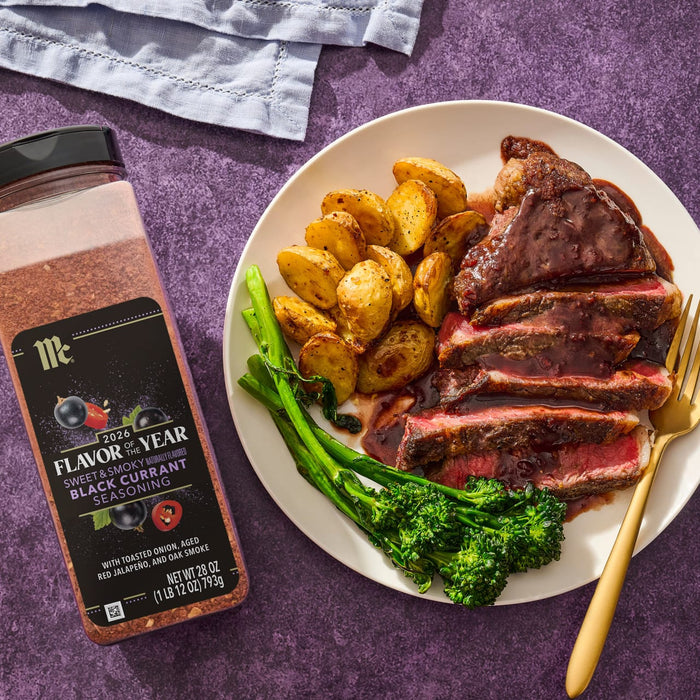 A bottle of McCormick 2026 Flavor of the Year Sweet &amp; Smoky Naturally Flavored Black Currant Seasoning sits next to a plate of steak, potatoes, and broccolini.
