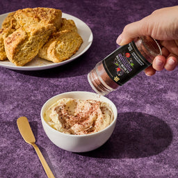 A hand sprinkles McCormick Flavor of the Year 2026 Sweet &amp; Smoky Black Currant Seasoning onto a bowl of cream, with scones and a butter knife in the background.