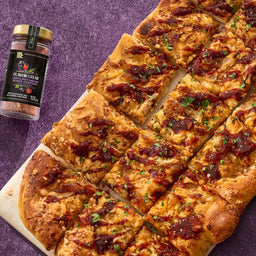 A McCormick Flavor of the Year 2026 Sweet &amp; Smoky Naturally Flavored Black Currant Seasoning jar sits next to a sliced flatbread pizza topped with caramelized onions and a dark sauce.