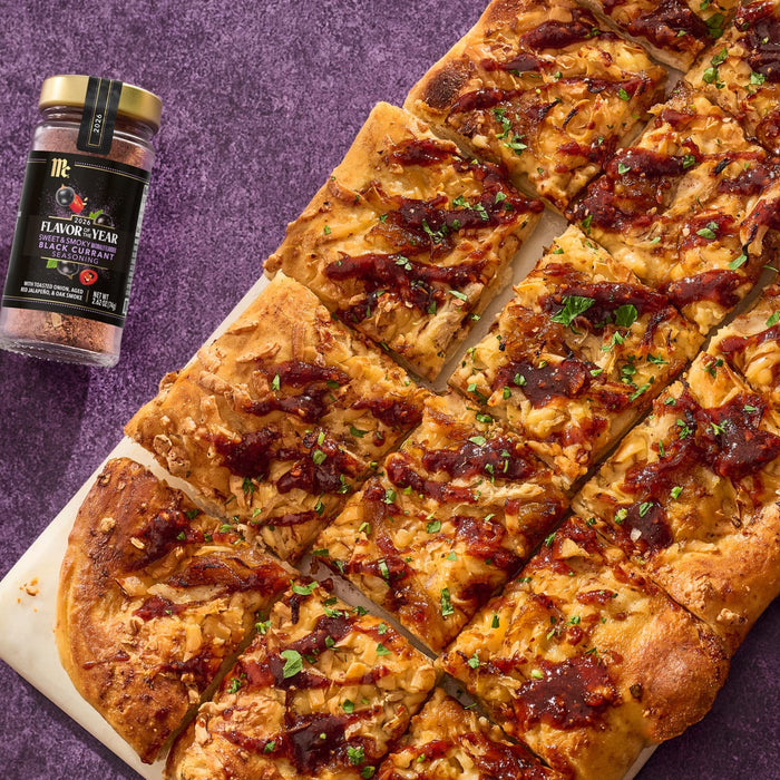 A McCormick Flavor of the Year 2026 Sweet &amp; Smoky Naturally Flavored Black Currant Seasoning jar sits next to a sliced flatbread pizza topped with caramelized onions and a dark sauce.