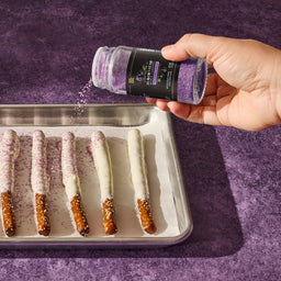 A hand sprinkles McCormick 2026 Flavor of the Year Black Currant Finishing Sugar onto white chocolate-covered pretzels.