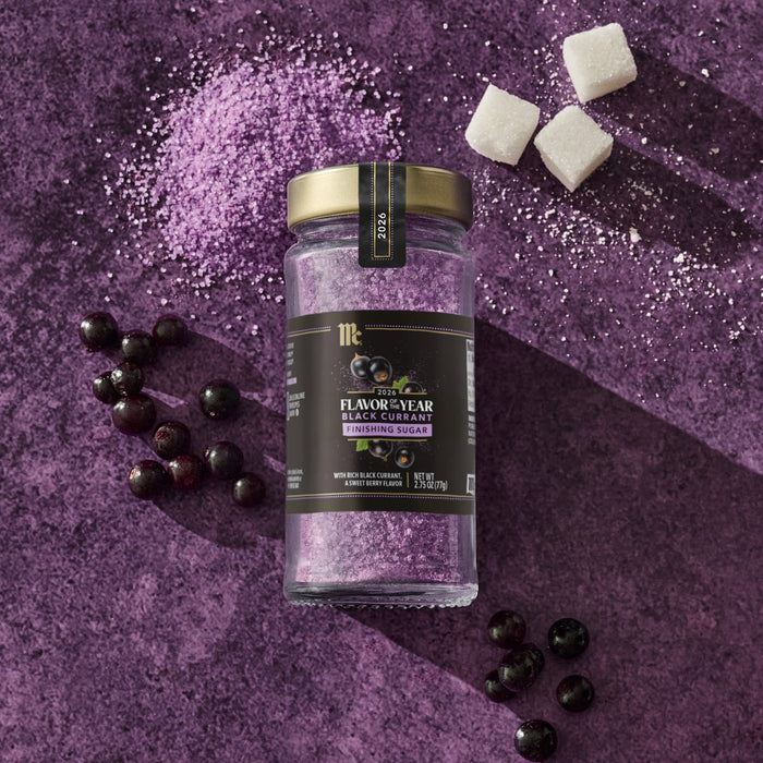 A jar of McCormick Flavor of the Year 2026 Black Currant Finishing Sugar is displayed with black currants, sugar cubes, and purple sugar crystals on a purple surface.