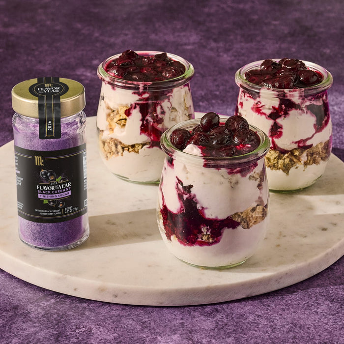 A jar of McCormick Flavor of the Year 2026 Black Currant Finishing Sugar sits next to three small glass jars filled with a layered dessert.