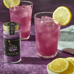 A jar of McCormick Flavor of the Year 2026 Black Currant Finishing Sugar is displayed next to two glasses of purple-colored drinks garnished with lemon slices.