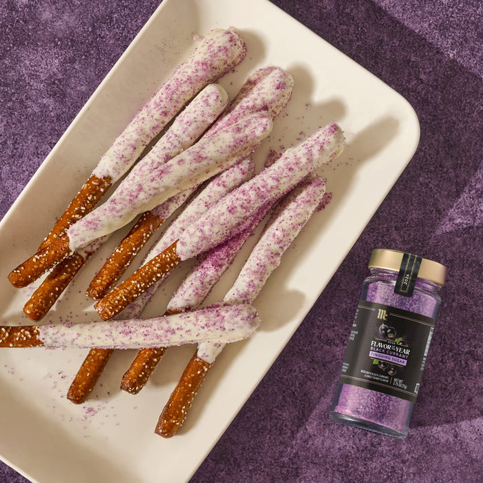 A white platter holds pretzel rods dipped in white chocolate and McCormick Flavor of the Year Black Currant Finishing Sugar, next to a jar of the finishing sugar.