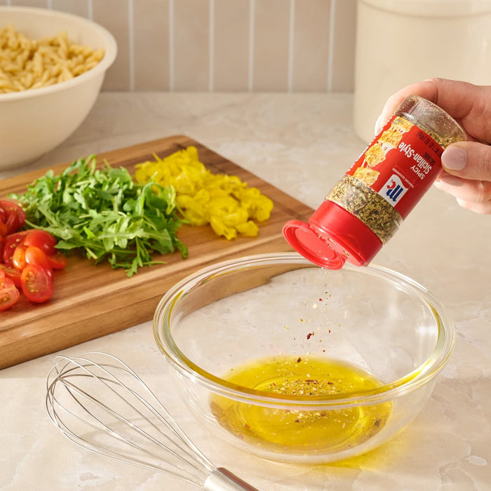 A hand shakes Mccormick Spicy Sicilian Style Seasoning, 1.62 Oz into a glass bowl of oil to create a dressing, with fresh pasta salad ingredients like tomatoes and arugula in the background.
