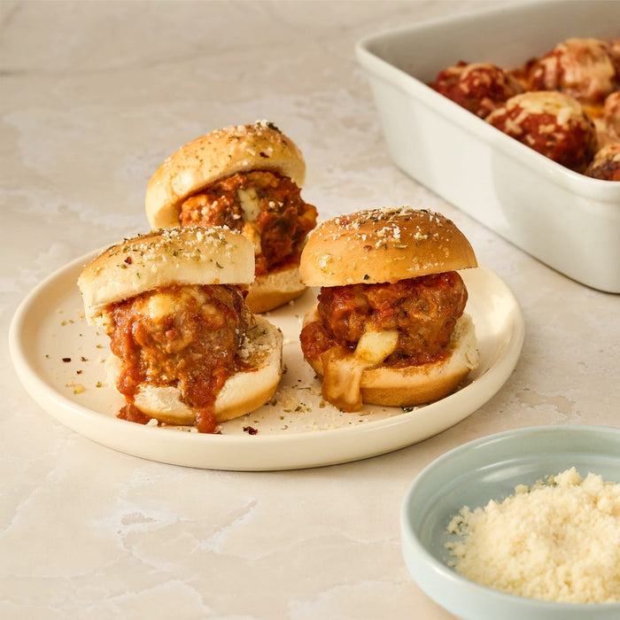 Three meatball sliders, made with Mccormick Spicy Sicilian Style Seasoning, 1.62 Oz, are arranged on a plate with a side of grated cheese.