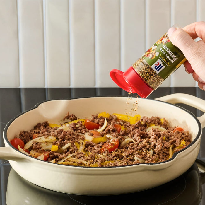 A hand sprinkles Mccormick Savory Herb Parmesan Seasoning, 2.5 Oz over a skillet of cooked ground beef, onions, and peppers.