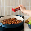 A hand sprinkles Mccormick Smoked Paprika And Garlic Seasoning, 3.12 Oz, over cooked ground meat in a blue and white pan, with taco ingredients like tortillas and lettuce nearby.