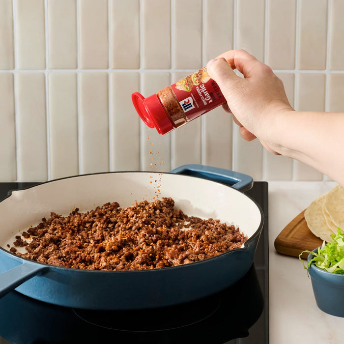 A hand sprinkles Mccormick Smoked Paprika And Garlic Seasoning, 3.12 Oz, over cooked ground meat in a blue and white pan, with taco ingredients like tortillas and lettuce nearby.