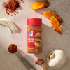 A bottle of Mccormick Smoked Paprika And Garlic Seasoning, 3.12 Oz, is displayed on a countertop surrounded by its fresh ingredients, including garlic, onion, turmeric, and paprika.