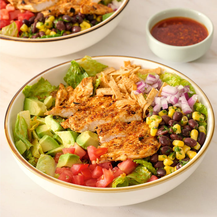 A close-up of a delicious-looking chicken salad in a white bowl, featuring chicken seasoned with Mccormick Smoked Paprika And Garlic Seasoning, 3.12 Oz, alongside fresh lettuce, avocado, and beans.