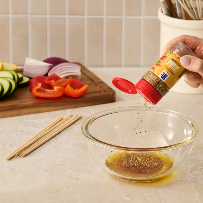 A hand pours Mccormick Rosemary Lemon Seasoning, 2.75 Oz, into a glass bowl of oil to create a marinade for fresh vegetables.