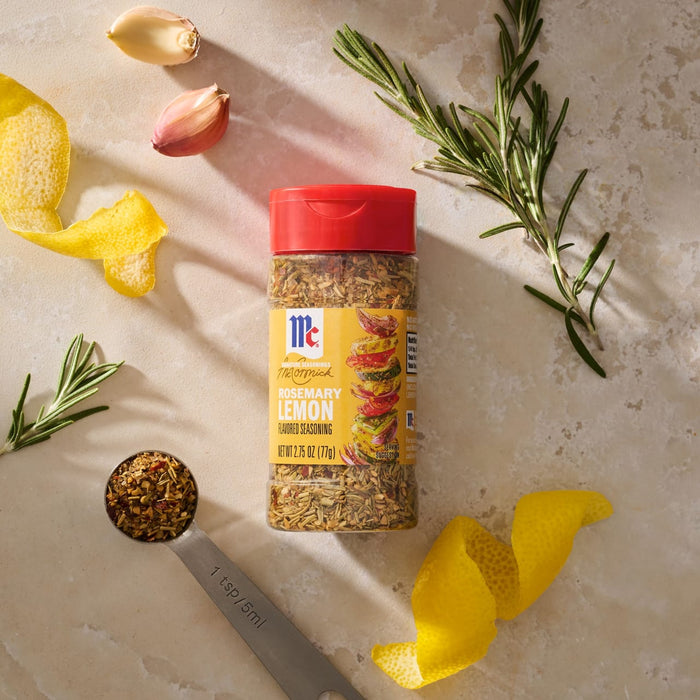 A bottle of Mccormick Rosemary Lemon Seasoning, 2.75 Oz, is artfully arranged on a light-colored surface with its fresh ingredients, including rosemary sprigs, lemon peels, garlic cloves, and a measuring spoon filled with the seasoning.