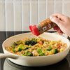 A hand sprinkles Mccormick Sesame Ginger Seasoning, 2.5 Oz, over a pan of vegetable fried rice on a stovetop to add a flavorful finishing touch.