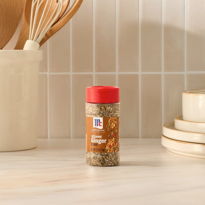 A bottle of Mccormick Sesame Ginger Seasoning, 2.5 Oz, is placed on a light-colored kitchen countertop, with kitchen utensils and plates in the background.