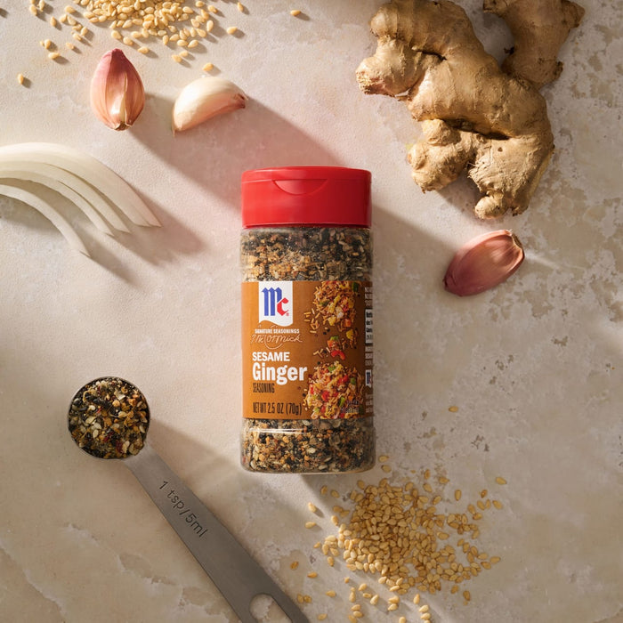 A bottle of Mccormick Sesame Ginger Seasoning, 2.5 Oz is arranged on a countertop with its fresh ingredients, including ginger, garlic, onion, and sesame seeds, to showcase its flavor profile.