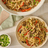 A plated serving of udon noodle stir-fry, made with Mccormick Sesame Ginger Seasoning, 2.5 Oz, sits next to the pan and a bowl of chopped green onions.
